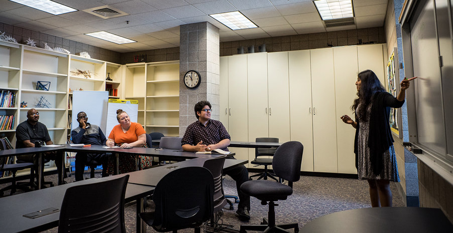 A female faculty member stands in the front of a conference room, presenting information to a group of colleagues.