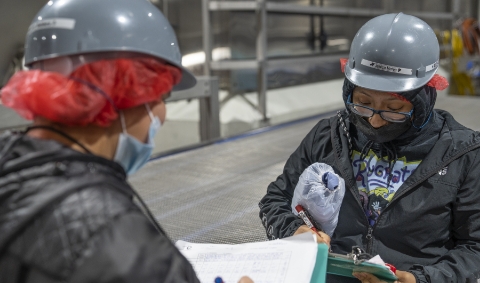  	Two Koch Foods employees in hard hats and face masks looking at paperwork on clipboards