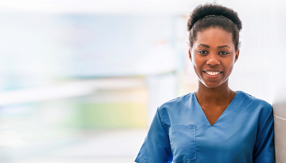 A young, female nurse in blue scrubs smiles as she walks with a female resident during her workday.