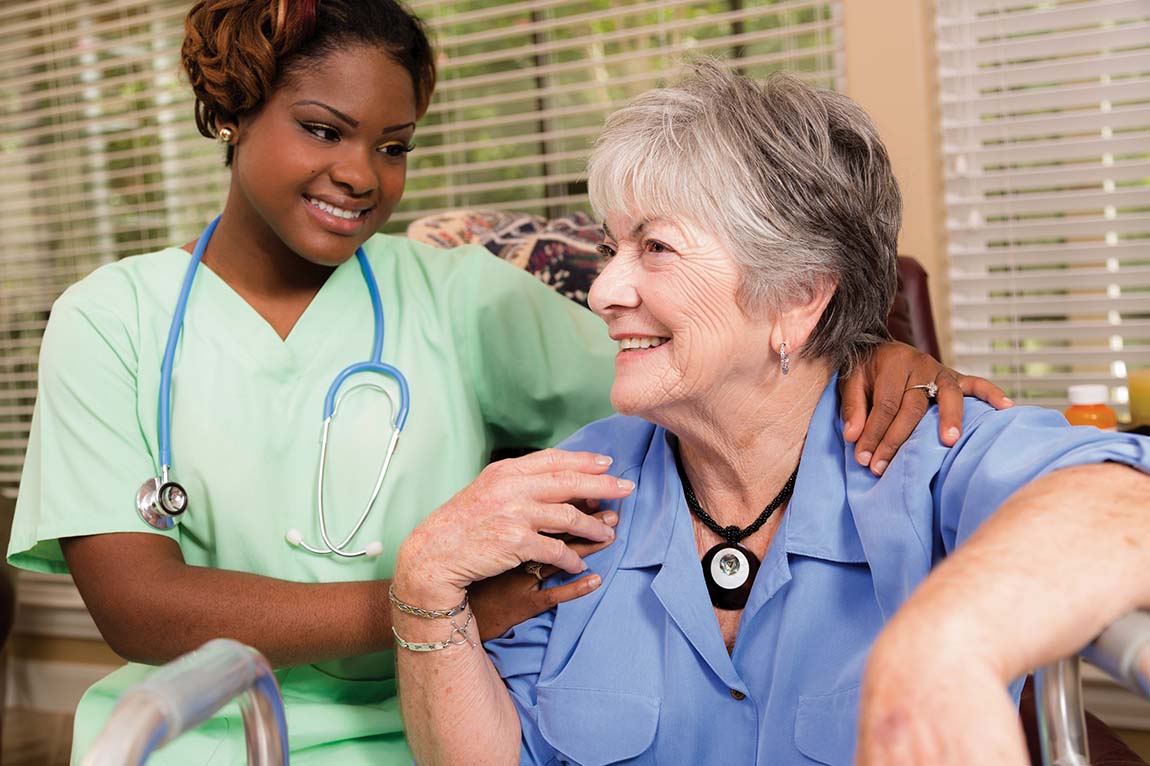 A smiling female STNA in lime-colored scrubs sits and talks to a female resident in her room.