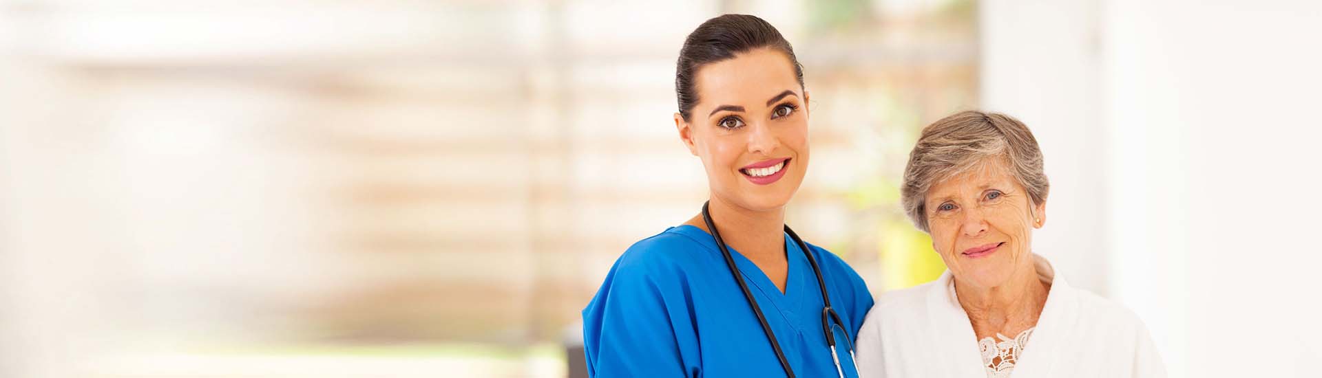 A young, female nurse in blue scrubs smiles as she walks with a female resident during her workday.