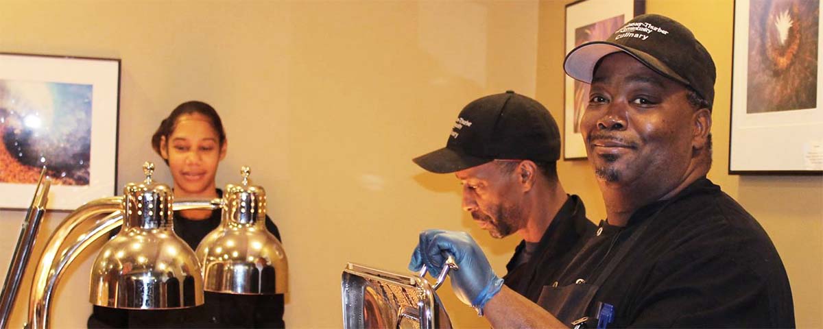 One female and two male staff members in black uniforms work at a food service area.
