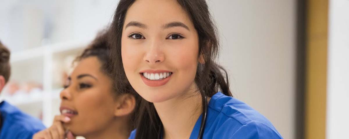 A young, female aide wearing blue scrubs smiles as she works with colleagues at station area.