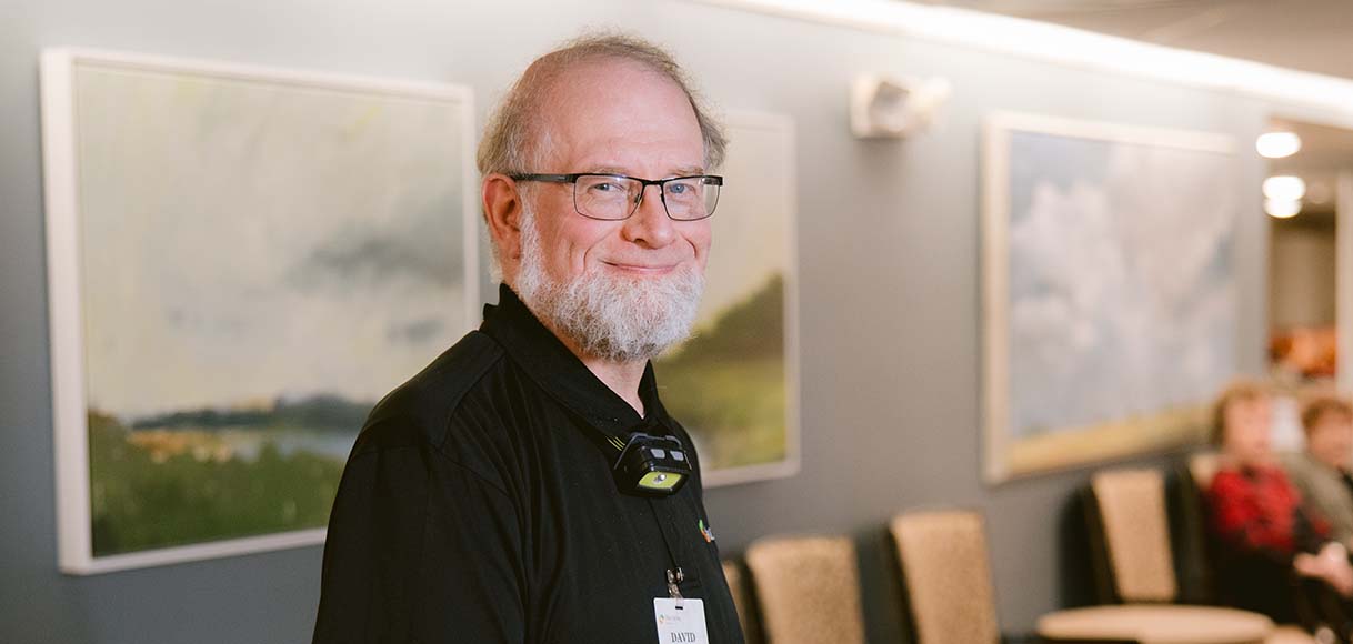 An older maintenance worker in a black Ohio Living polo shirt pauses to smile in a facility meeting room during his busy workday.