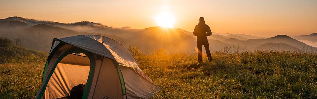 A hiker stands outside their tent and watches the sun set