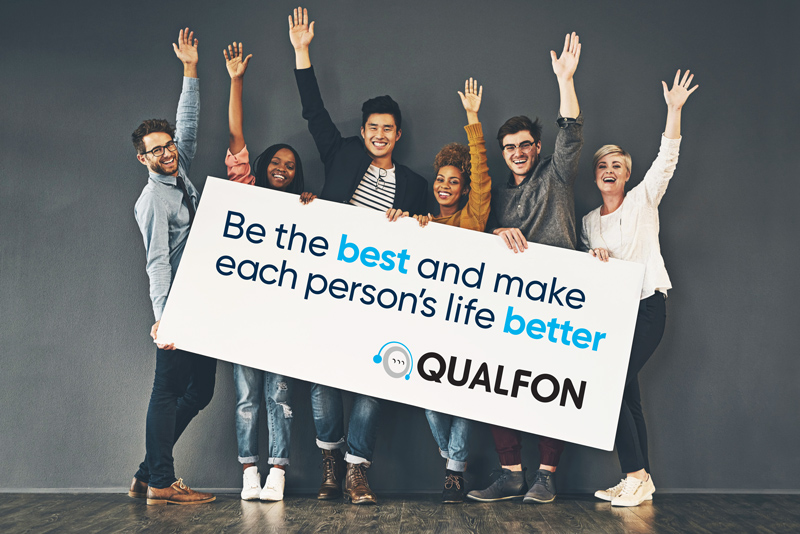 A group of people smiling and waving as they hold a sign with the Qualfon mission statement: Be the best and make each person's life better